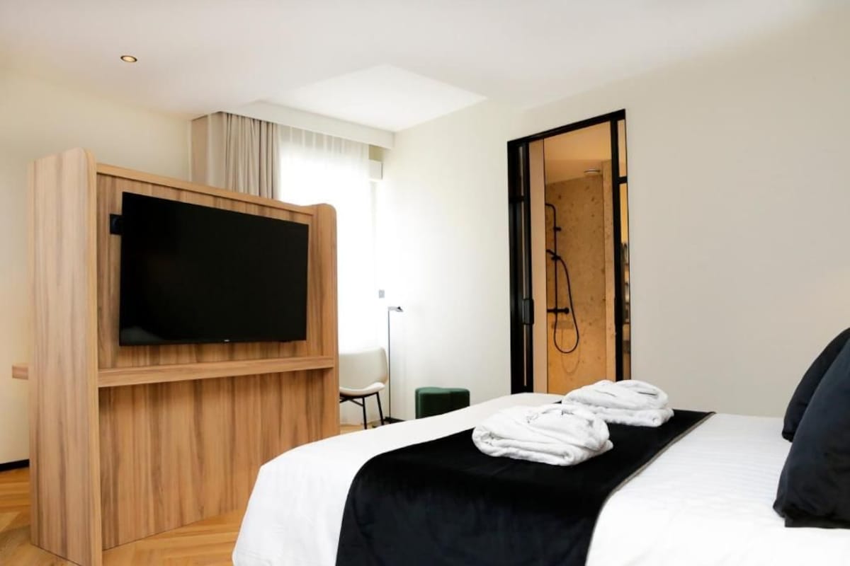 A hotel room with a bed, a TV mounted on a wooden panel, a shower area, and a green chair.
