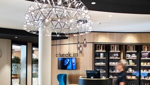A modern lobby with a check-in counter, a blue armchair, a round black table with a decorative item, and a large geometric light fixture.