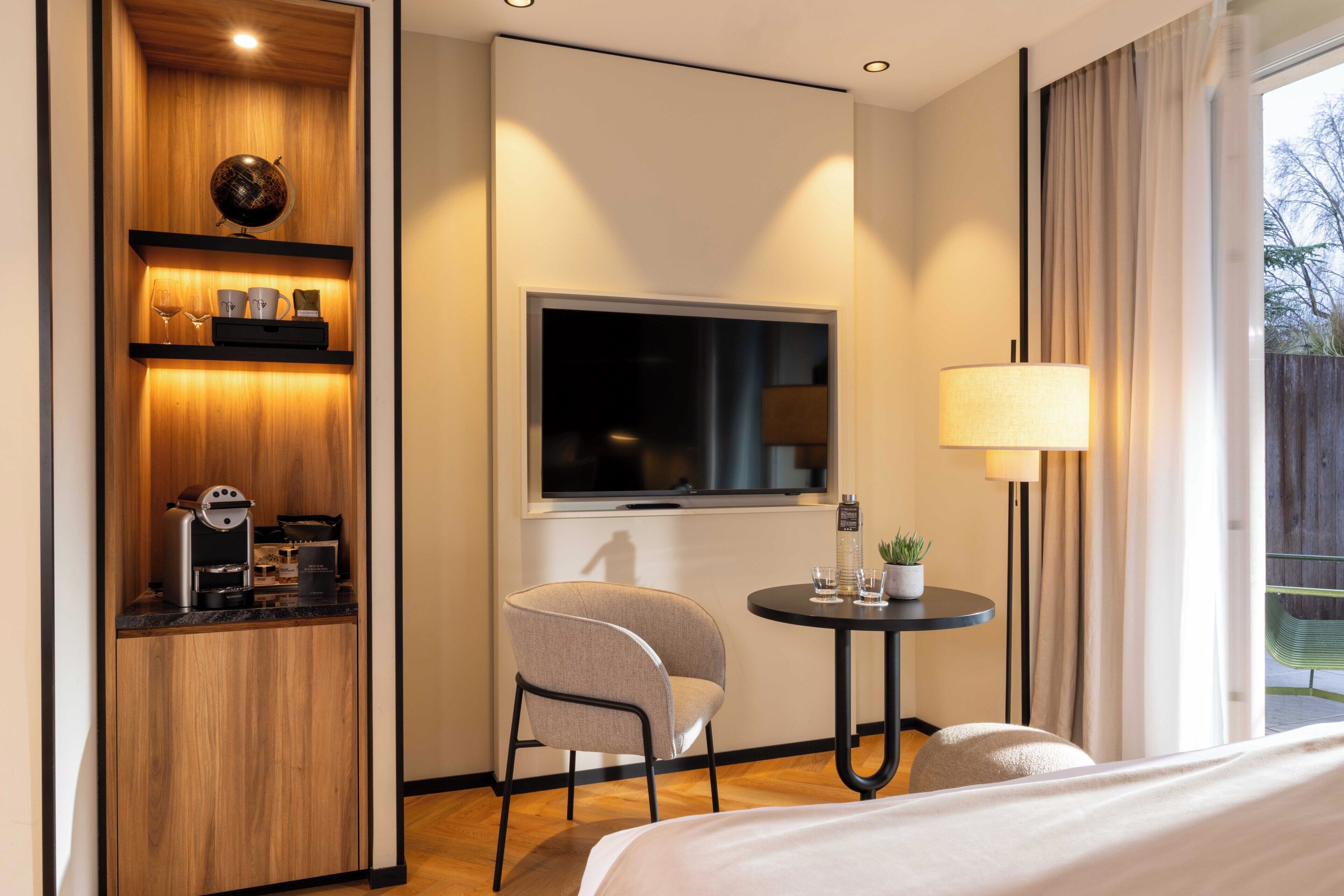 Quarto de hotel com cama, escrivaninha com luminária, cadeira, televisão e minibar.