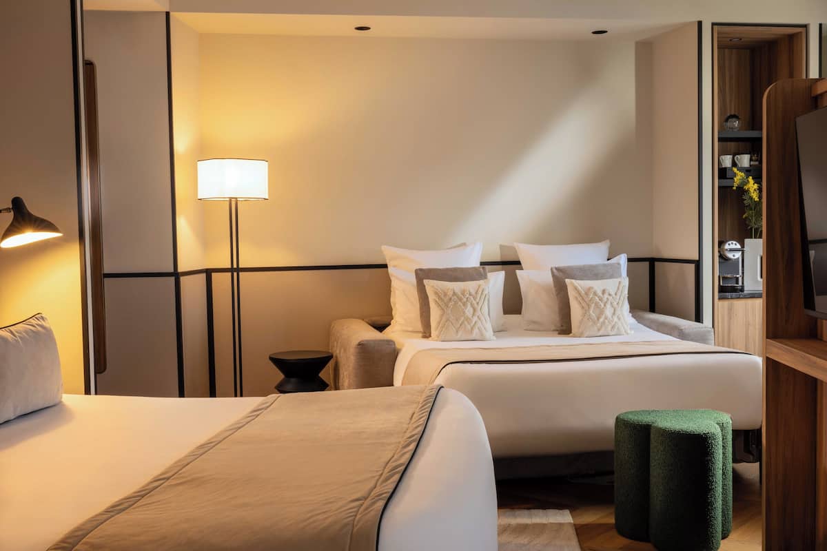 A hotel room with two beds, a floor lamp, a green ottoman, and a TV.