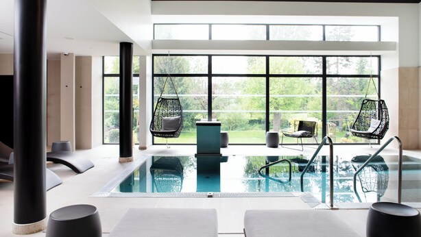A modern indoor swimming pool with clear glass walls, black hanging chairs, and a view of greenery outside.