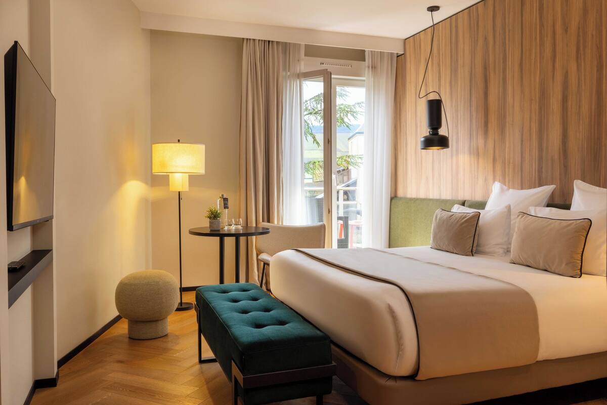 A hotel room with a bed, a small table with a lamp, a round ottoman, and a TV mounted on the wall.