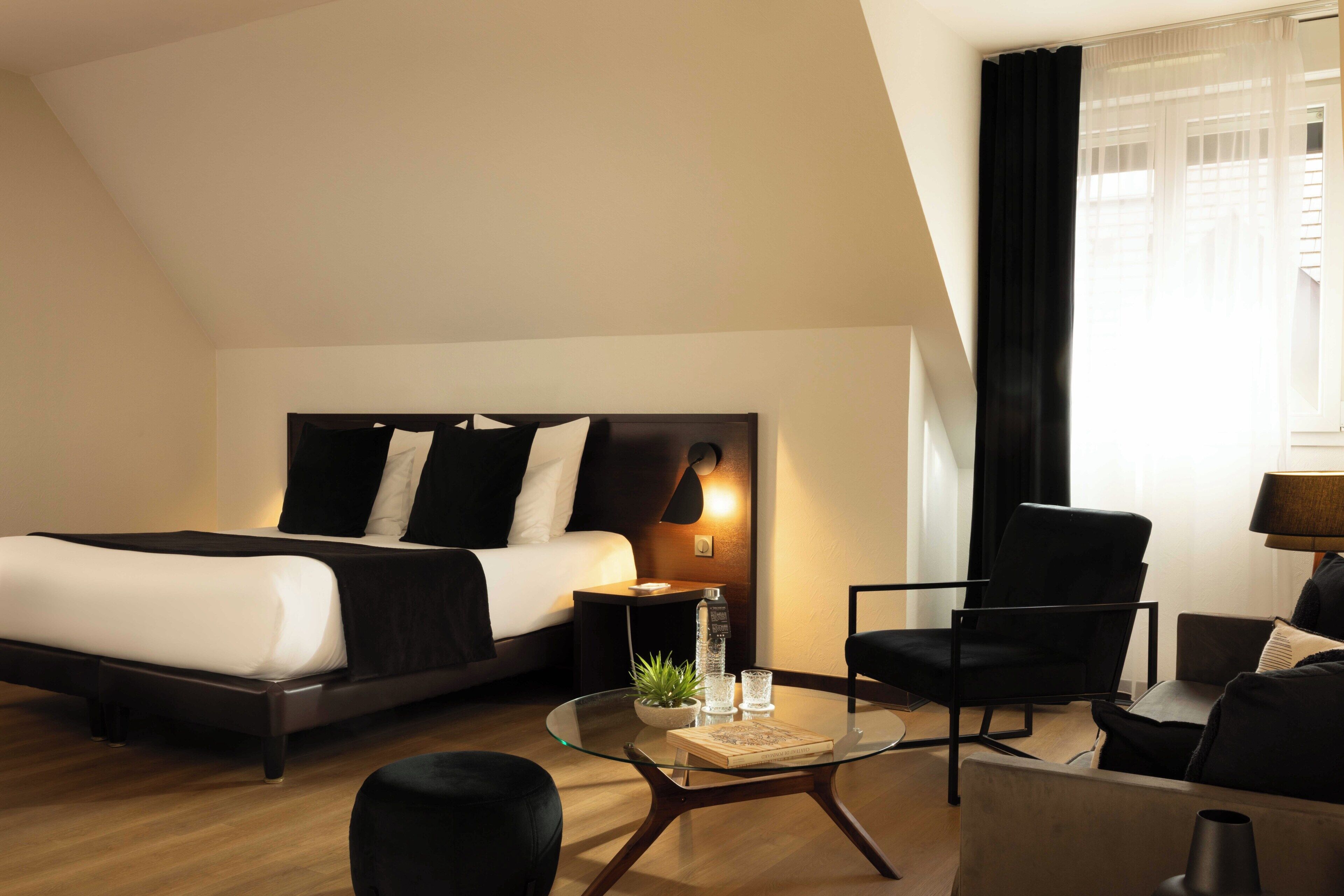 A modern hotel room with a bed, a glass coffee table, a chair, a sofa, and a nightstand with a lamp.