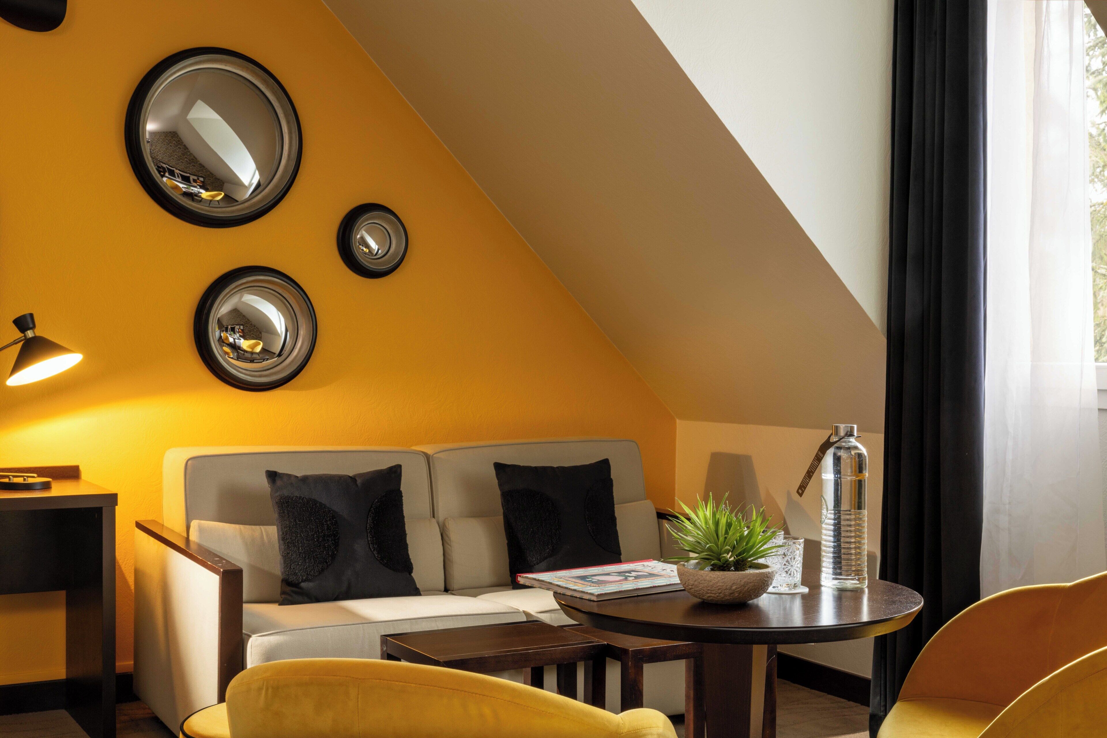 Una habitación moderna con sofá, un sillón amarillo, una mesa redonda con una planta y una botella de agua, y tres espejos montados en la pared.