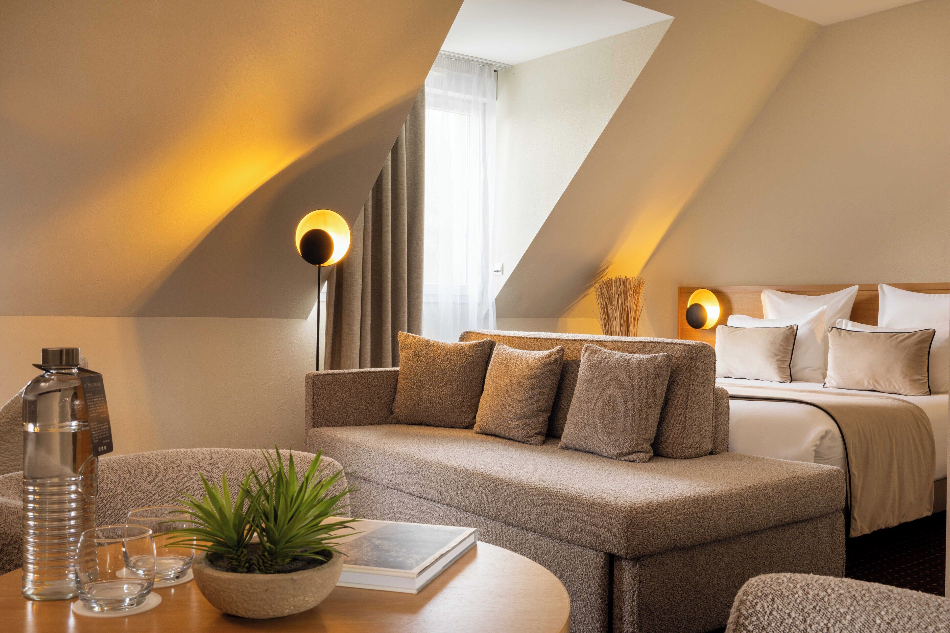 A modern hotel room with a sofa, a bedside table with a plant, a lamp, and a bed with pillows.
