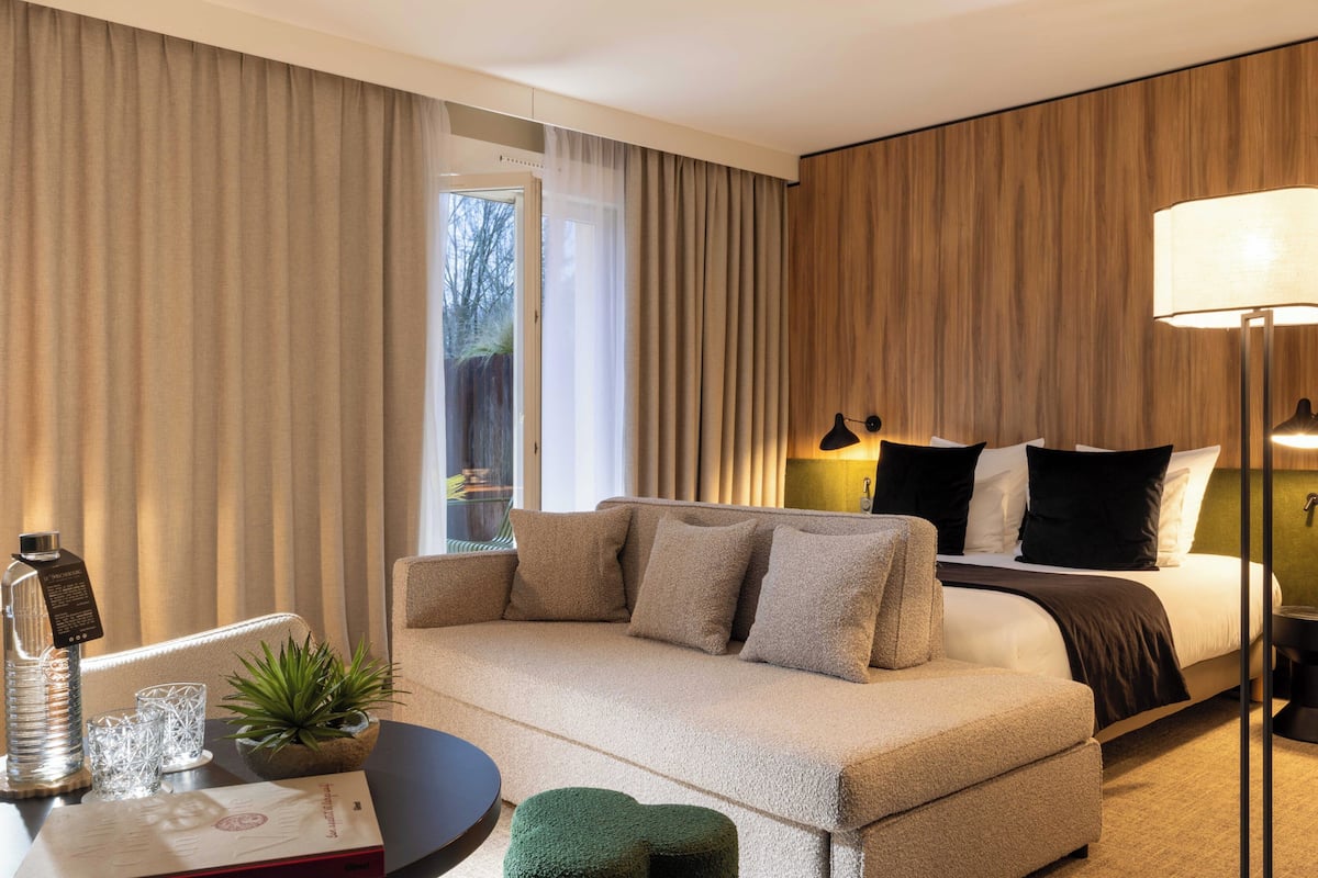 A hotel room with a bed, a sofa, a small table with a lamp, and a window with curtains.