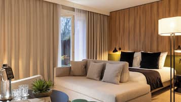 Una habitación de hotel con cama, sofá, una mesita con lámpara y ventana con cortinas.