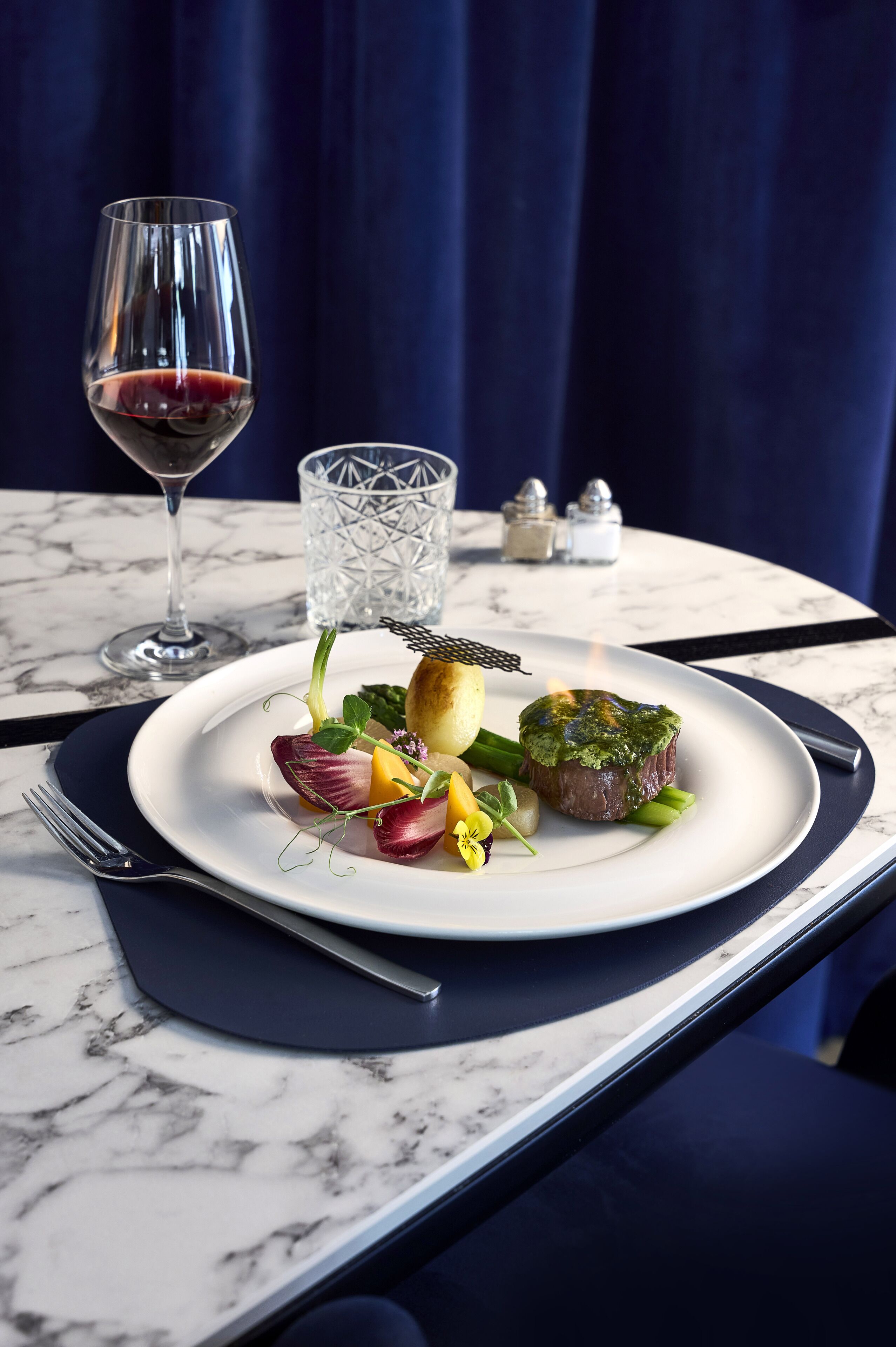 A dining table with a white plate of food, a glass of red wine, and a clear glass.