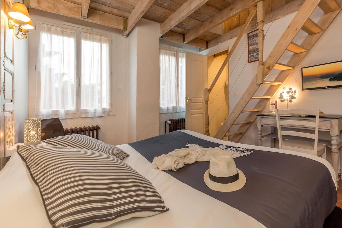 Une chambre à coucher avec un lit, des oreillers à rayures, un chapeau et une vue sur un escalier menant à l’étage supérieur.