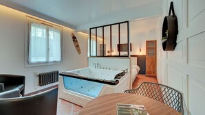 Een moderne badkamer met een badkuip, een douche en een toilet. Er is een raam met gordijnen, een handdoekhouder en een klein tafeltje met een boek.