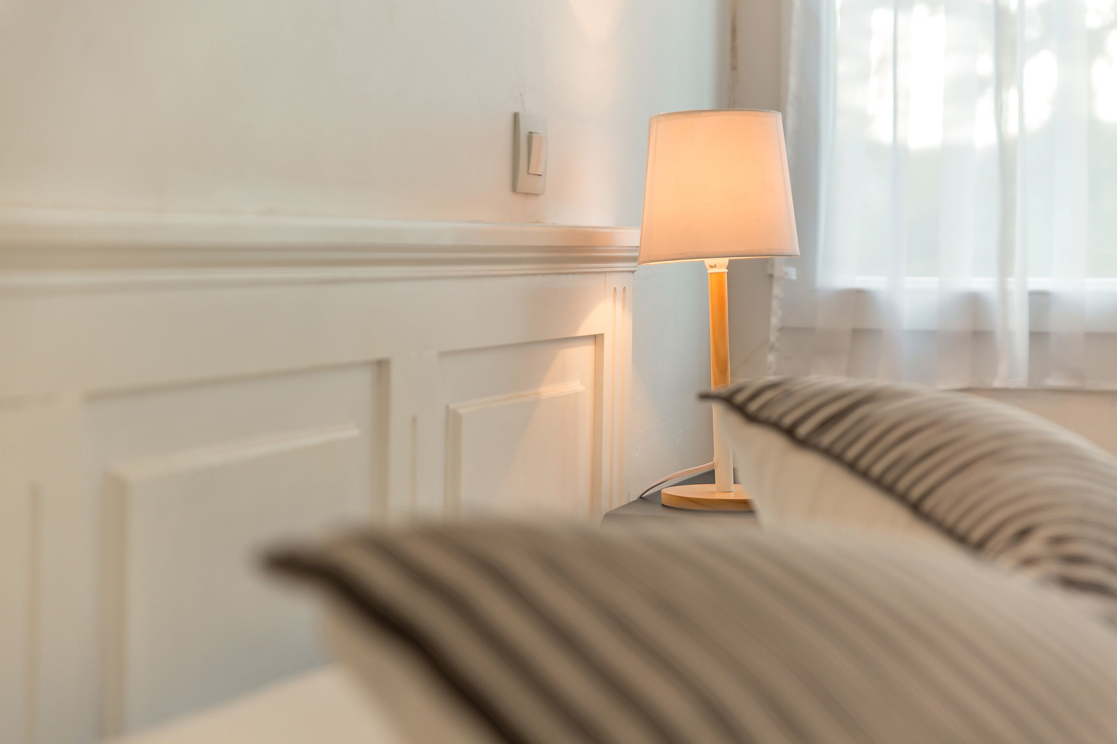 Une chambre avec une table de chevet, une lampe et une fenêtre ornée de rideaux légers.