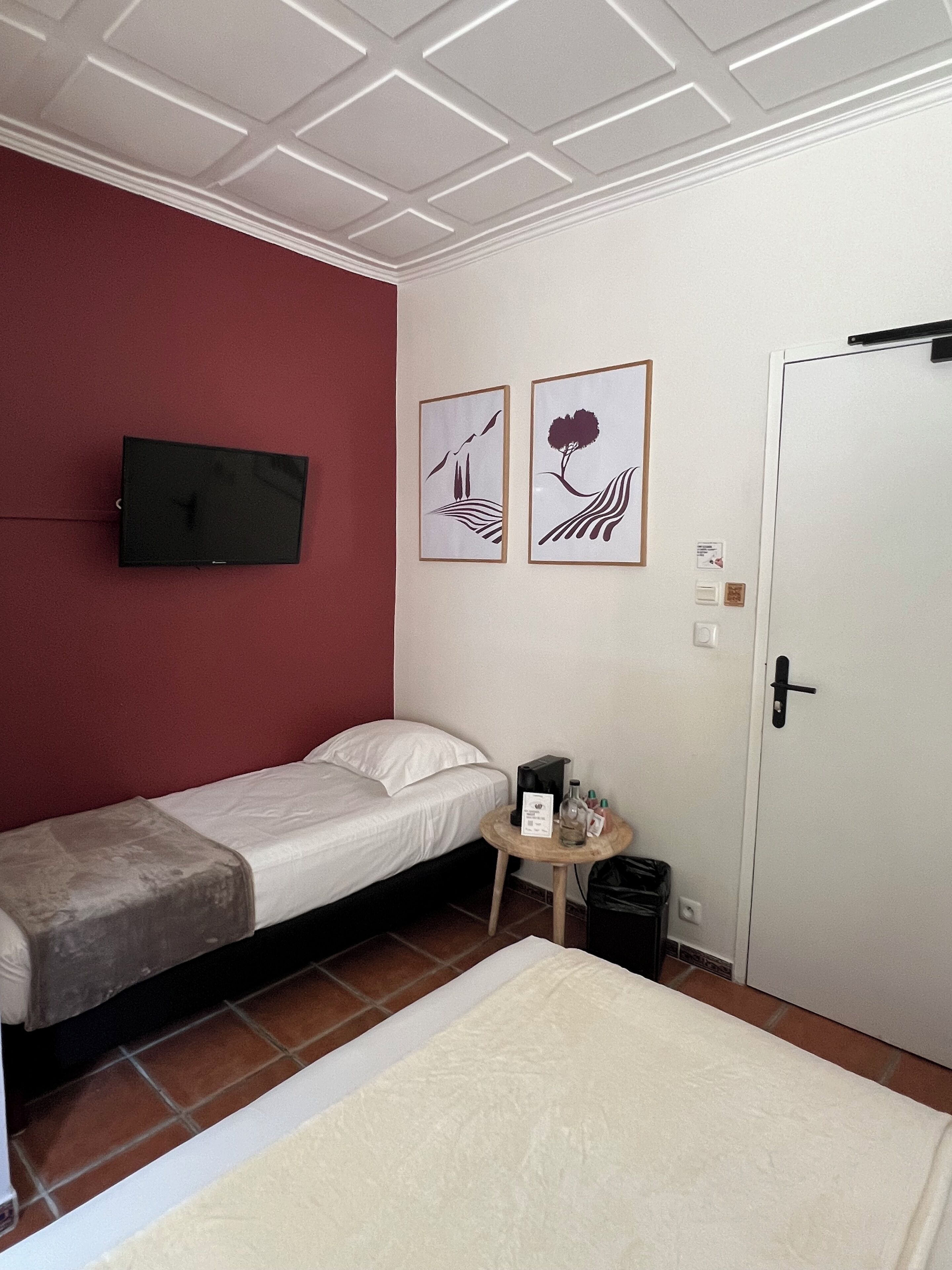 Une petite pièce, bien rangée, avec un lit simple, une petite table sur laquelle se trouvent une bouteille et une télécommande, et une porte.