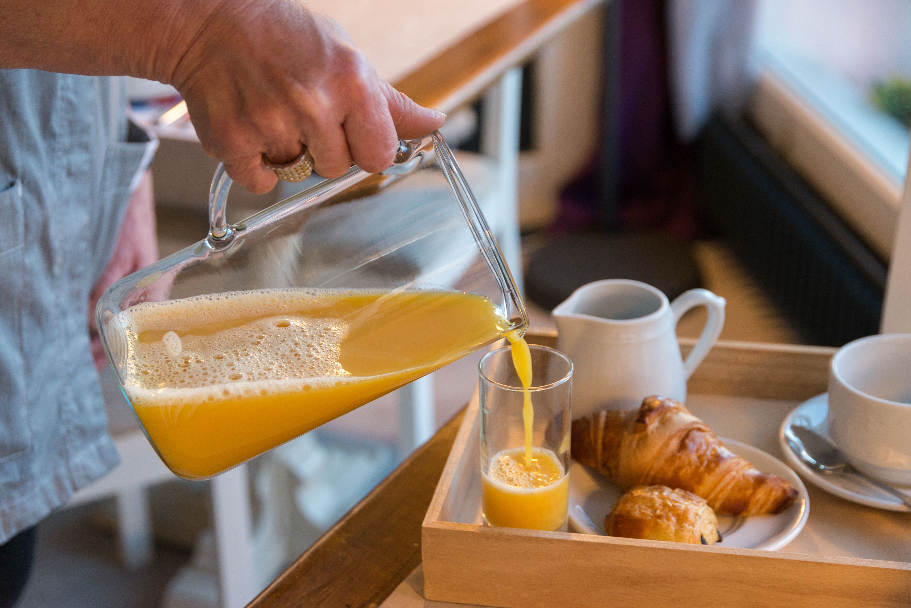 A person pouring orange juice into a glass on a tray with croissants and cups.