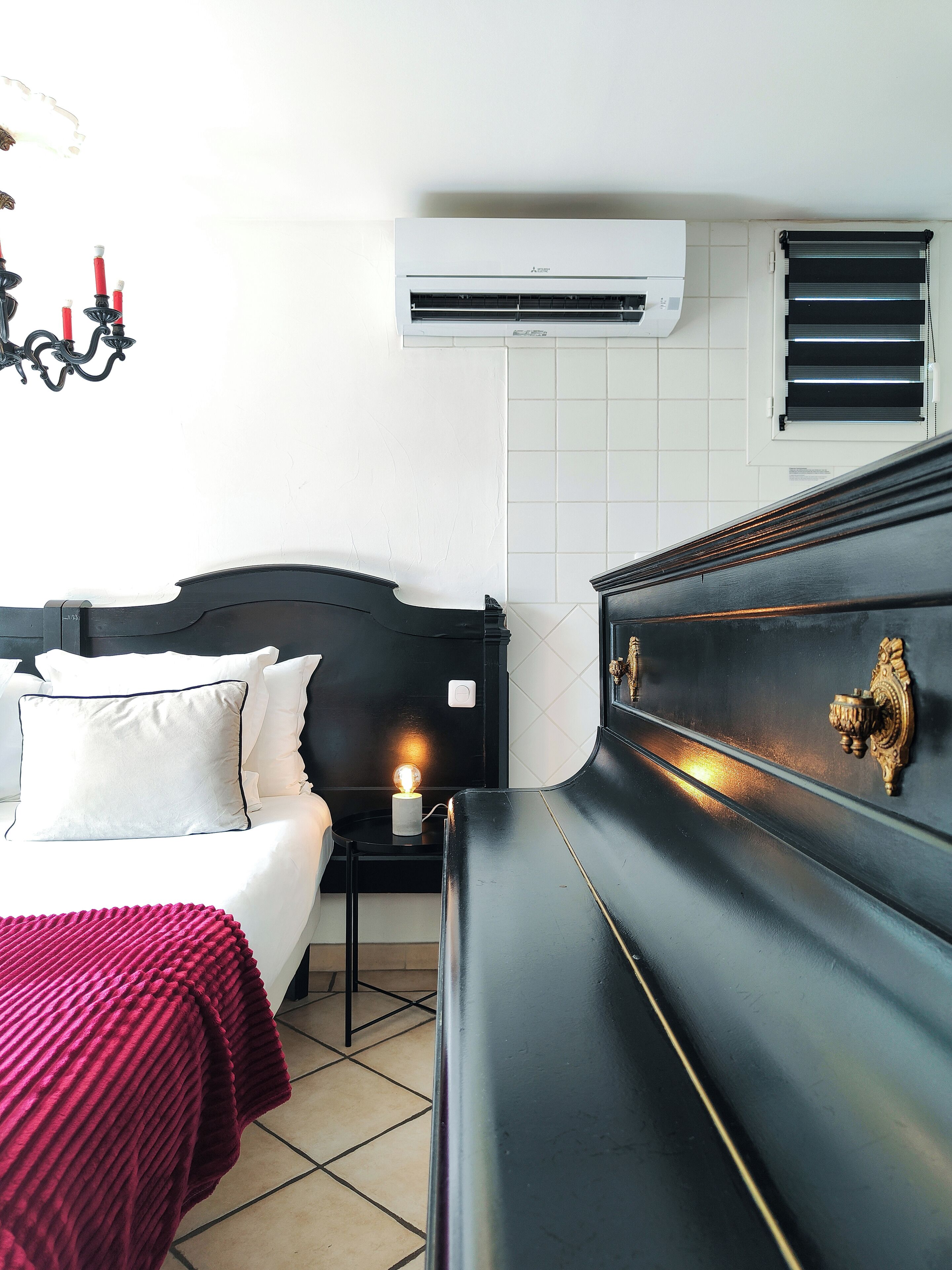 A hotel room with a bed, bedside table, lamp, and an air conditioning unit.