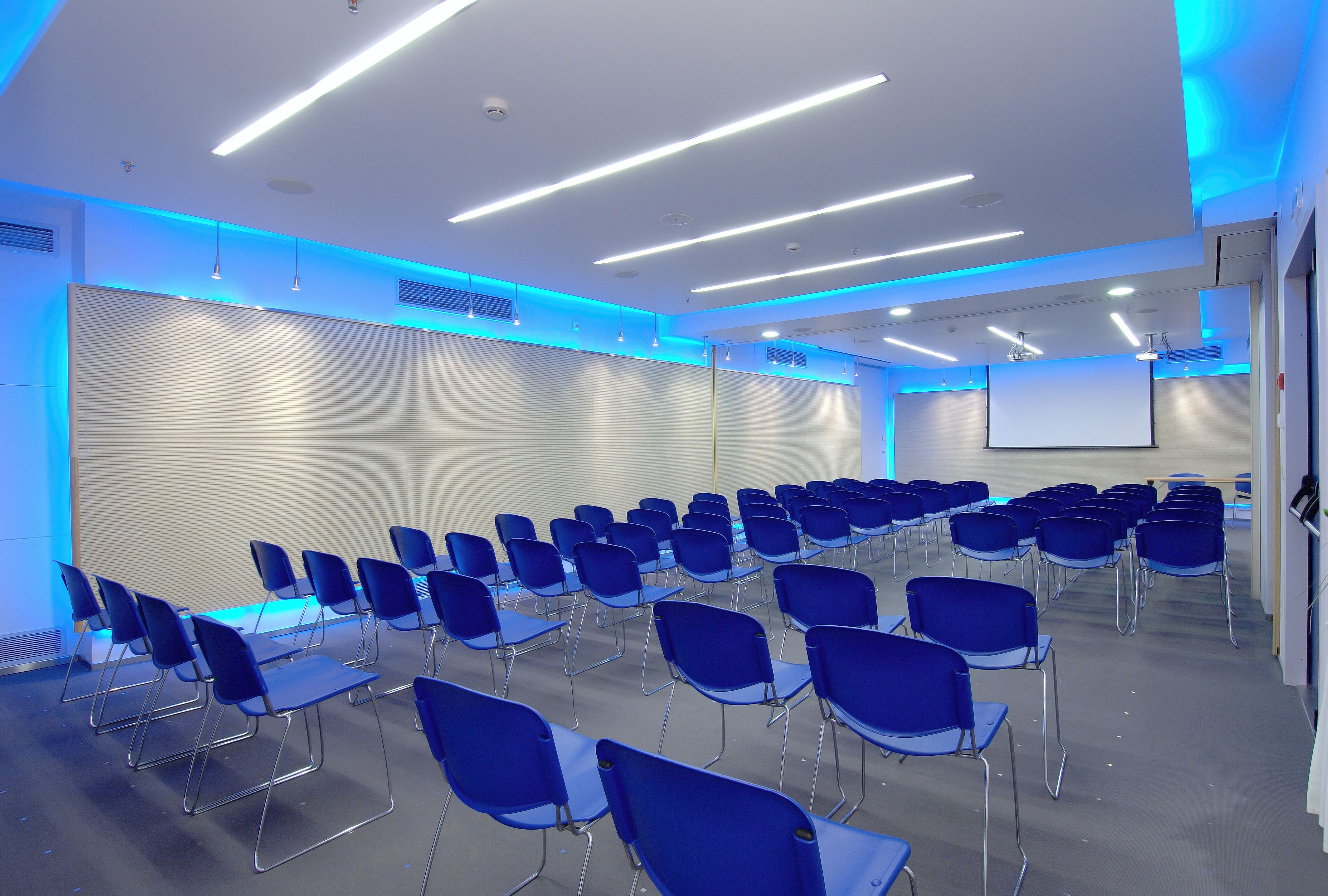 A conference room with rows of blue chairs, a white projection screen, and modern lighting.