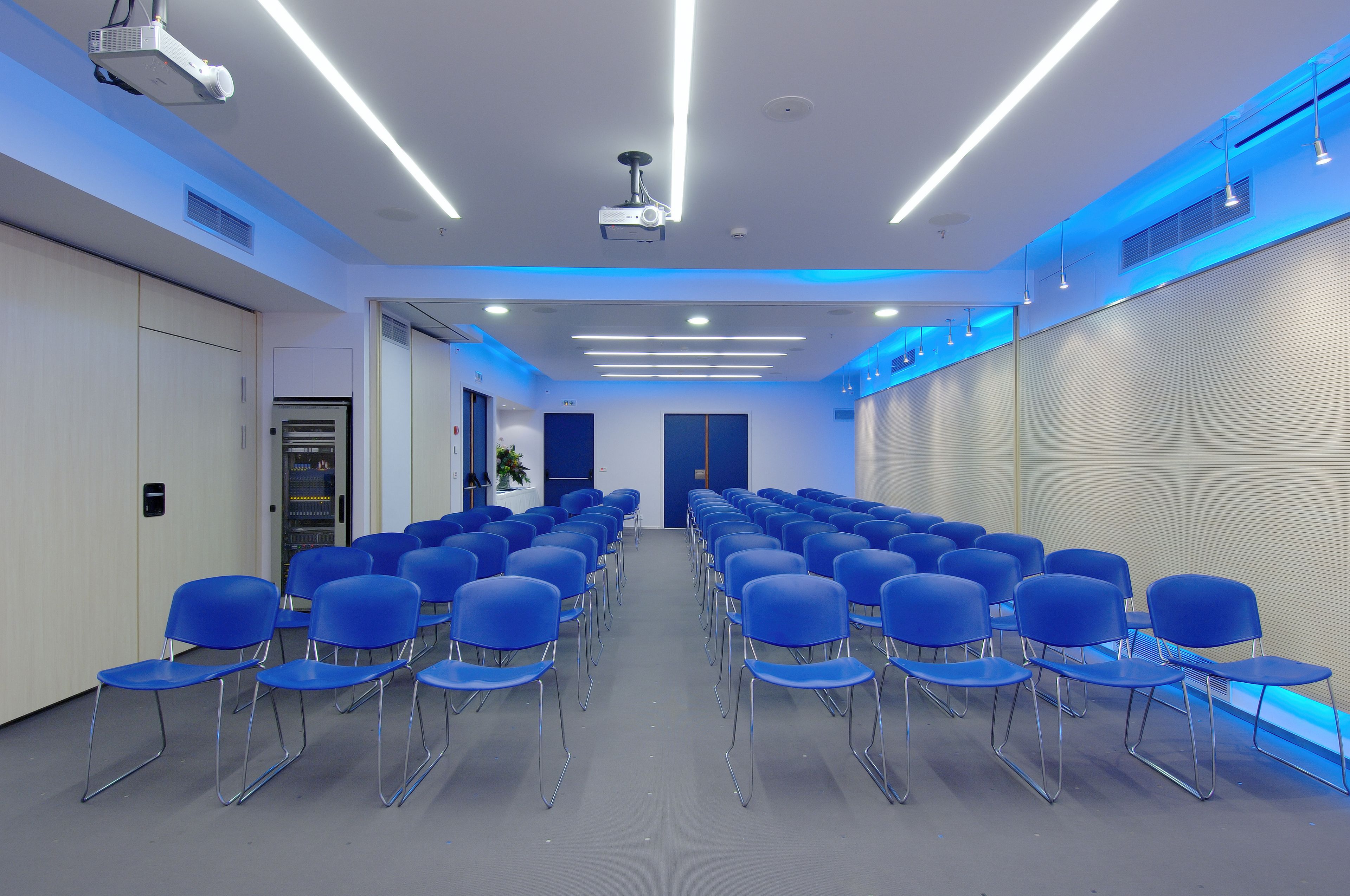 A conference room with rows of blue chairs, a projector, and ceiling lights.
