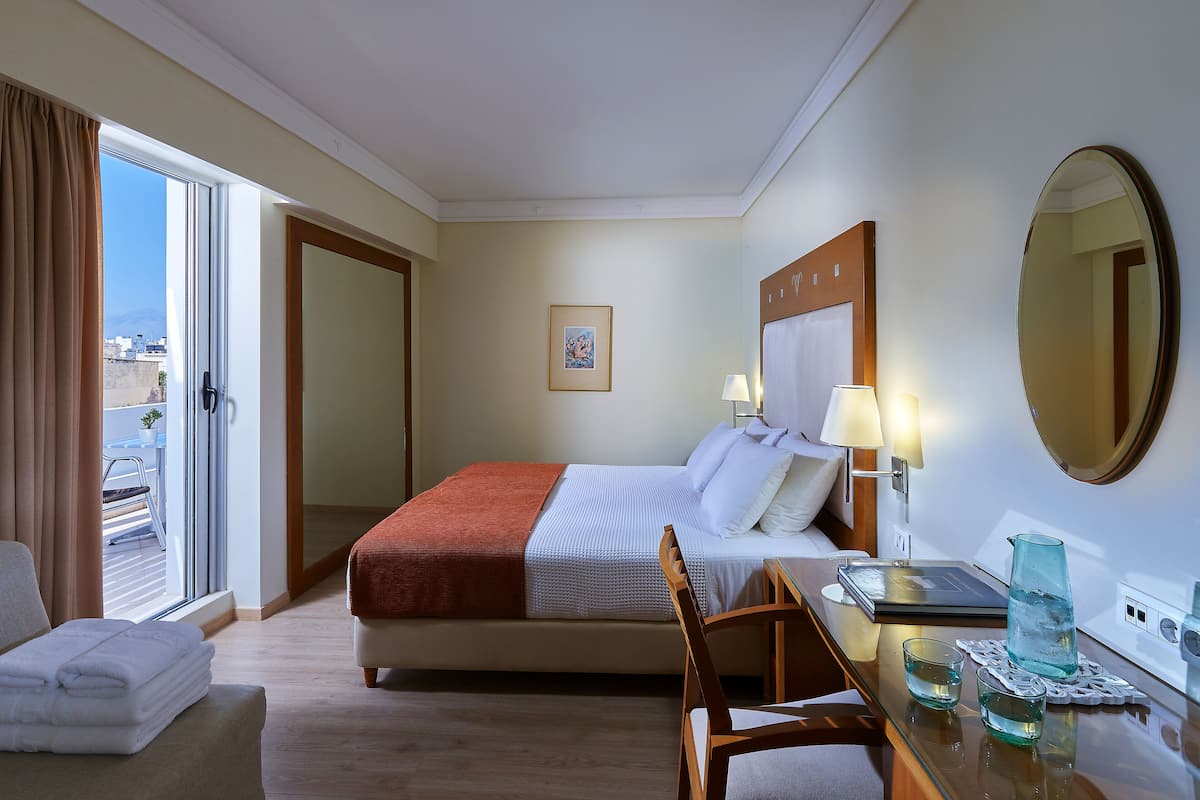A hotel room with a bed, a desk with a laptop, a chair, a table with a glass pitcher, and a view of the city through the window.