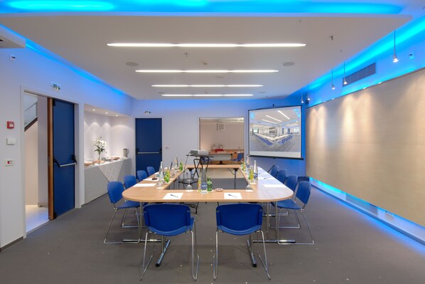 Una sala de conferencias moderna con una larga mesa, sillas azules y una pantalla de proyector.