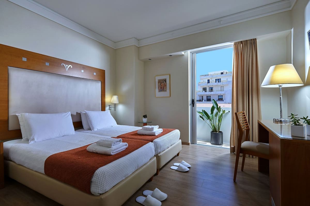 A hotel room with two beds, a desk, a chair, a potted plant, and a view of the city.