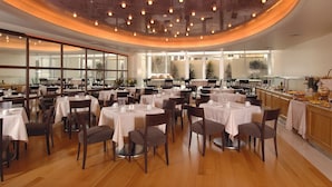 A restaurant with multiple tables set for dining, a kitchen area with food preparation visible, and large windows allowing natural light. - Atrion Hotel (Heraklion)
