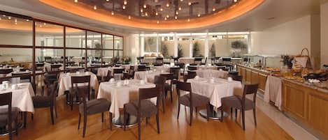 A restaurant with multiple tables set for dining, a kitchen area with food preparation visible, and large windows allowing natural light.