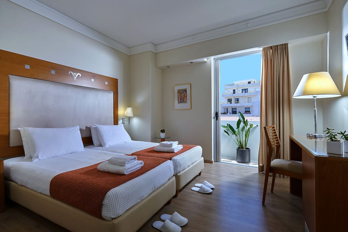A hotel room with a large bed, a desk, a chair, and a balcony with a view of buildings.