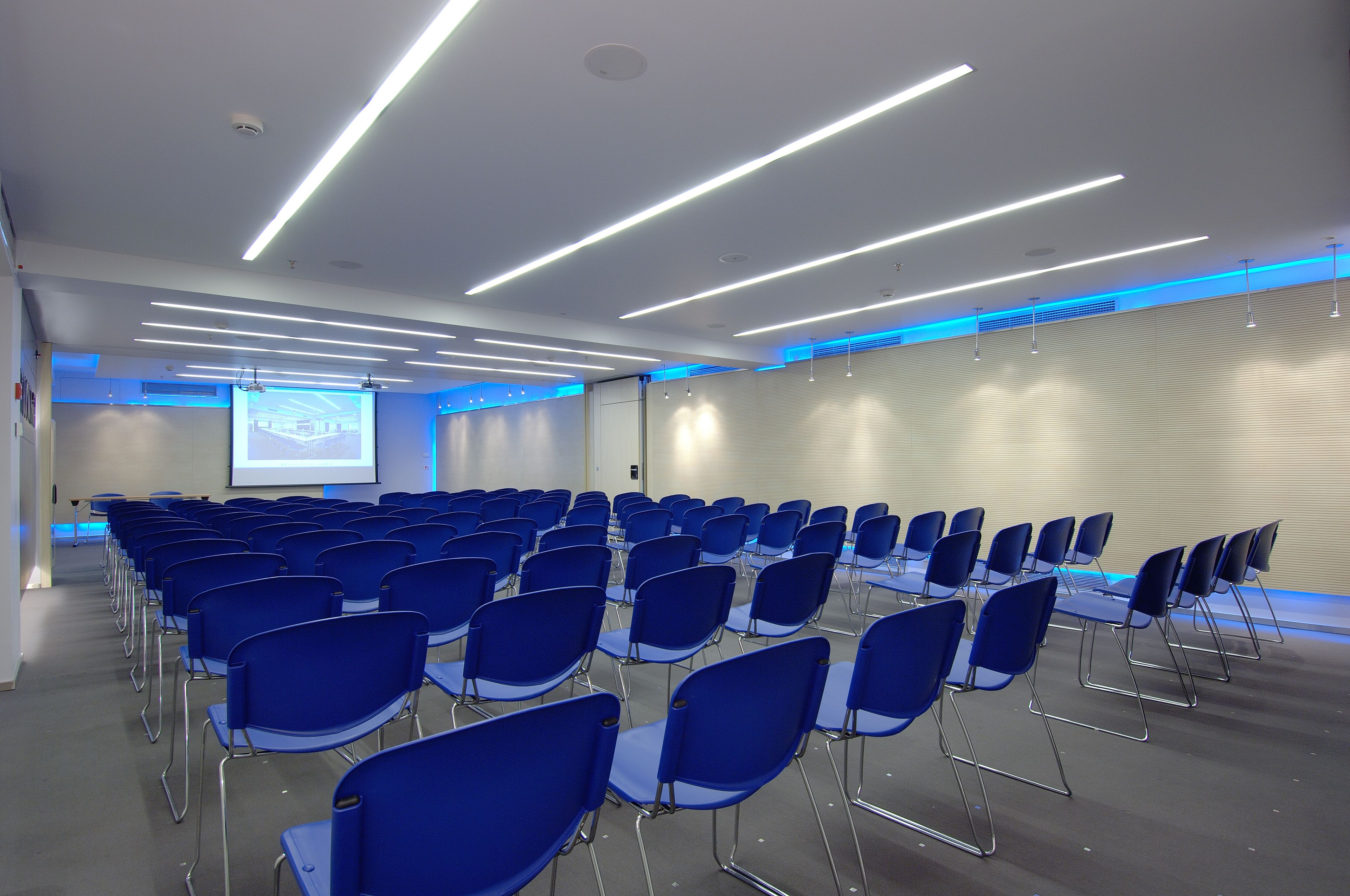 Un salón de conferencias con filas de sillas azules, una pantalla de proyector e iluminación moderna.
