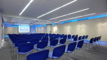 Un salón de conferencias con filas de sillas azules, una pantalla de proyector e iluminación moderna.