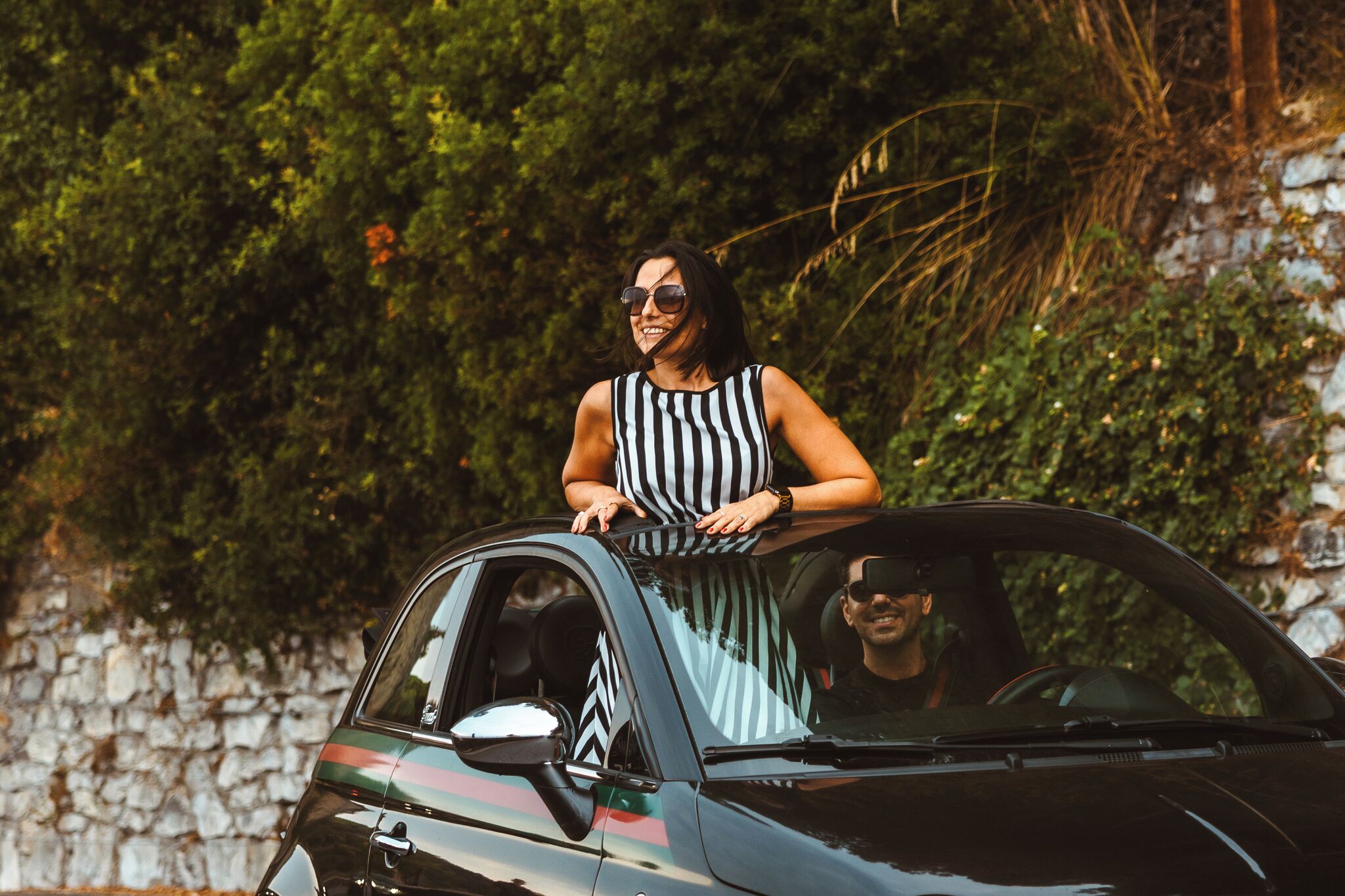 A person standing on the roof of a car with a stone wall in the background.