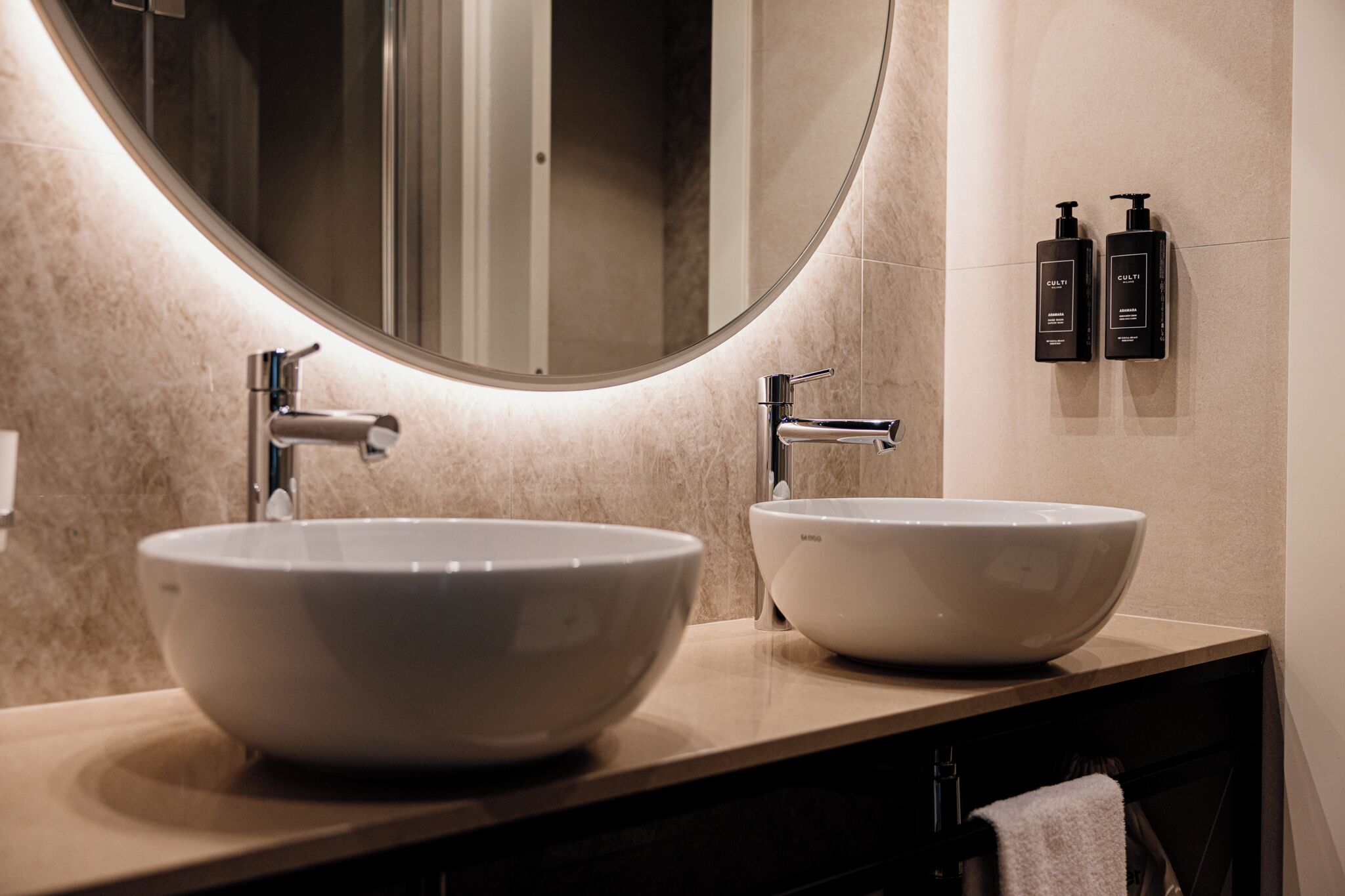 A bathroom with two large white vessel sinks, a mirror, and two black toiletries on the wall.