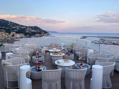A rooftop terrace with wicker furniture and round tables, overlooking a coastal area with buildings and a pier.
