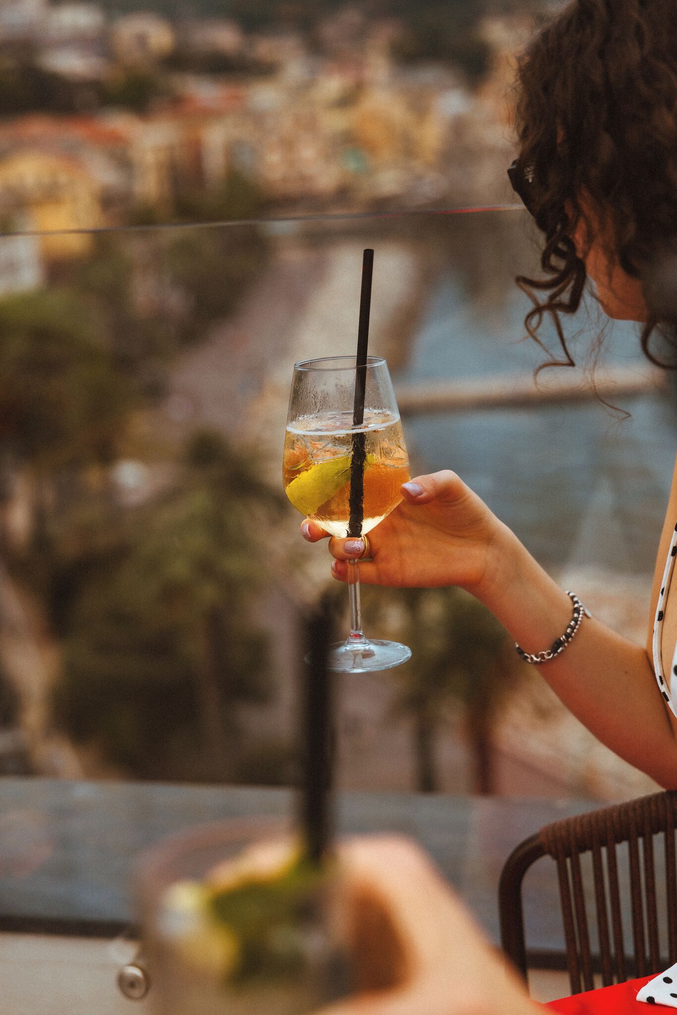 A person holding a glass with a straw, with a blurred background of a building and a body of water.