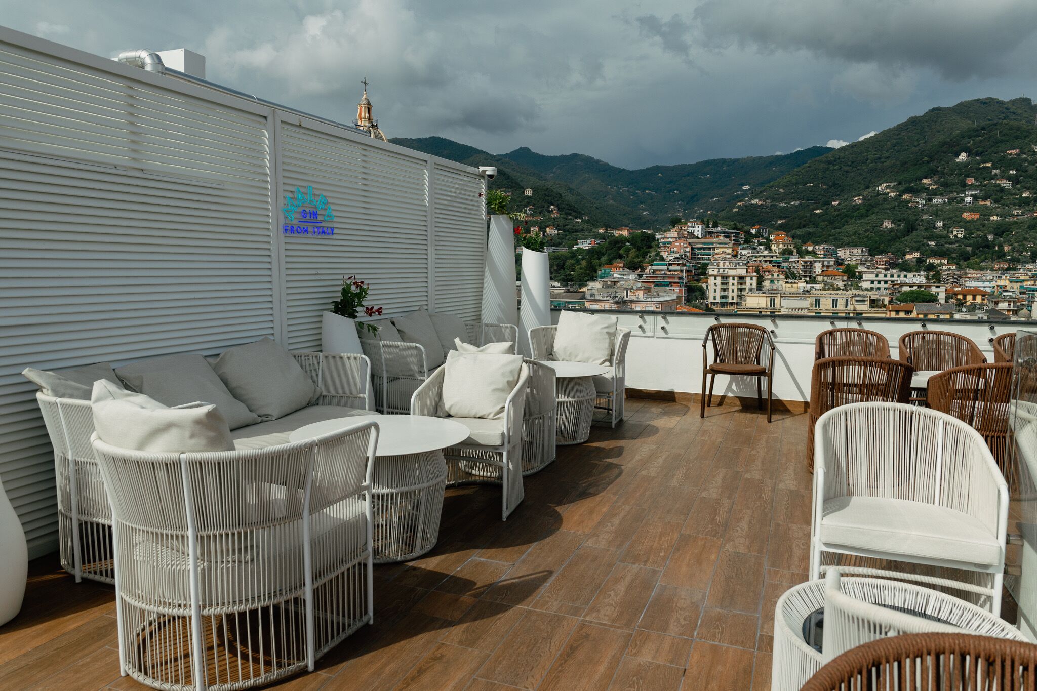 Une terrasse sur le toit, meublée de mobilier en osier, offrant une vue sur une ville côtière, et un panneau indiquant qu'on y sert une cuisine italienne.