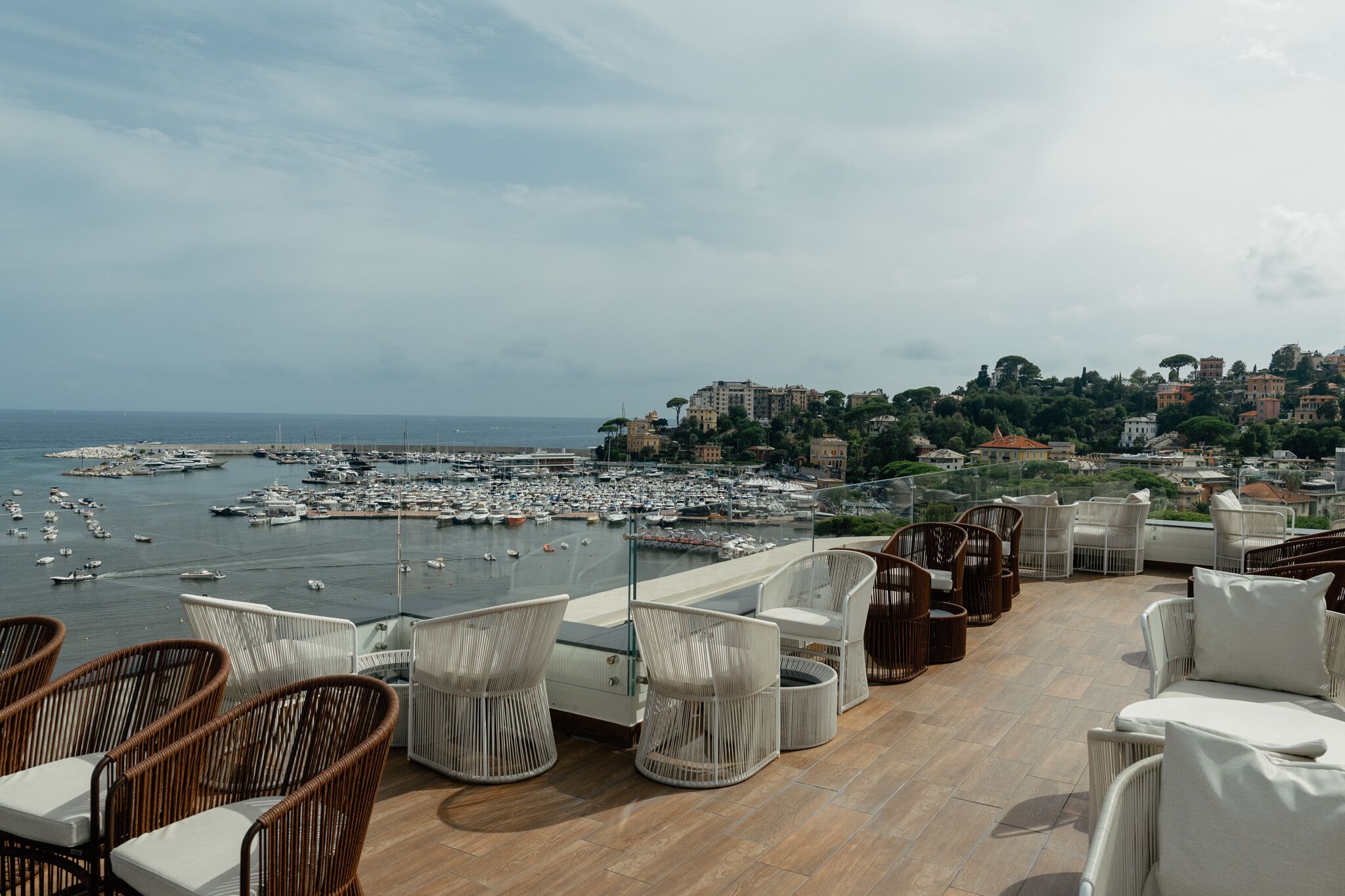 A terrace with wicker furniture overlooking a marina and a coastal town.