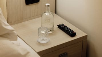 A wooden bedside table with a remote control, a glass bottle, and a glass.