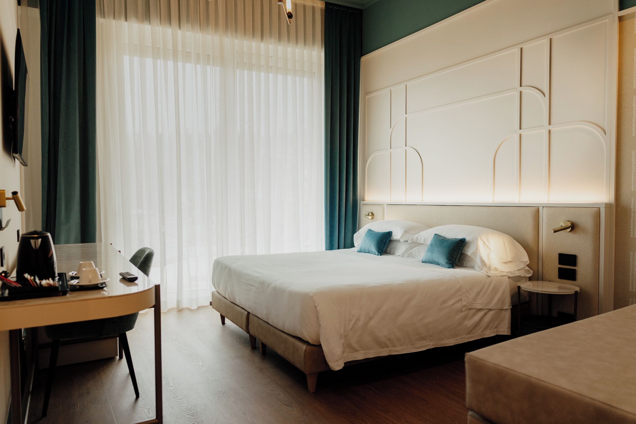 A hotel room with a bed, bedside tables, a desk with a chair, and a window with curtains.