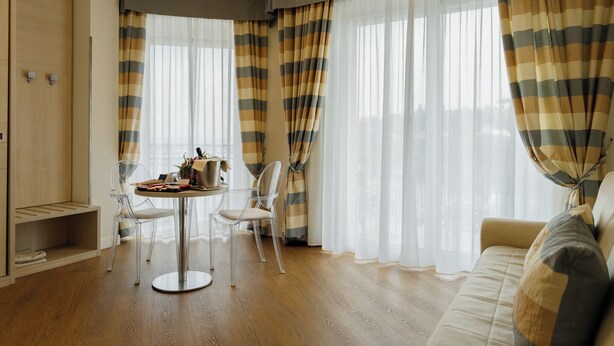 Een kamer met houten vloer, een ronde tafel met stoelen, een bank en grote ramen met gestreepte gordijnen.