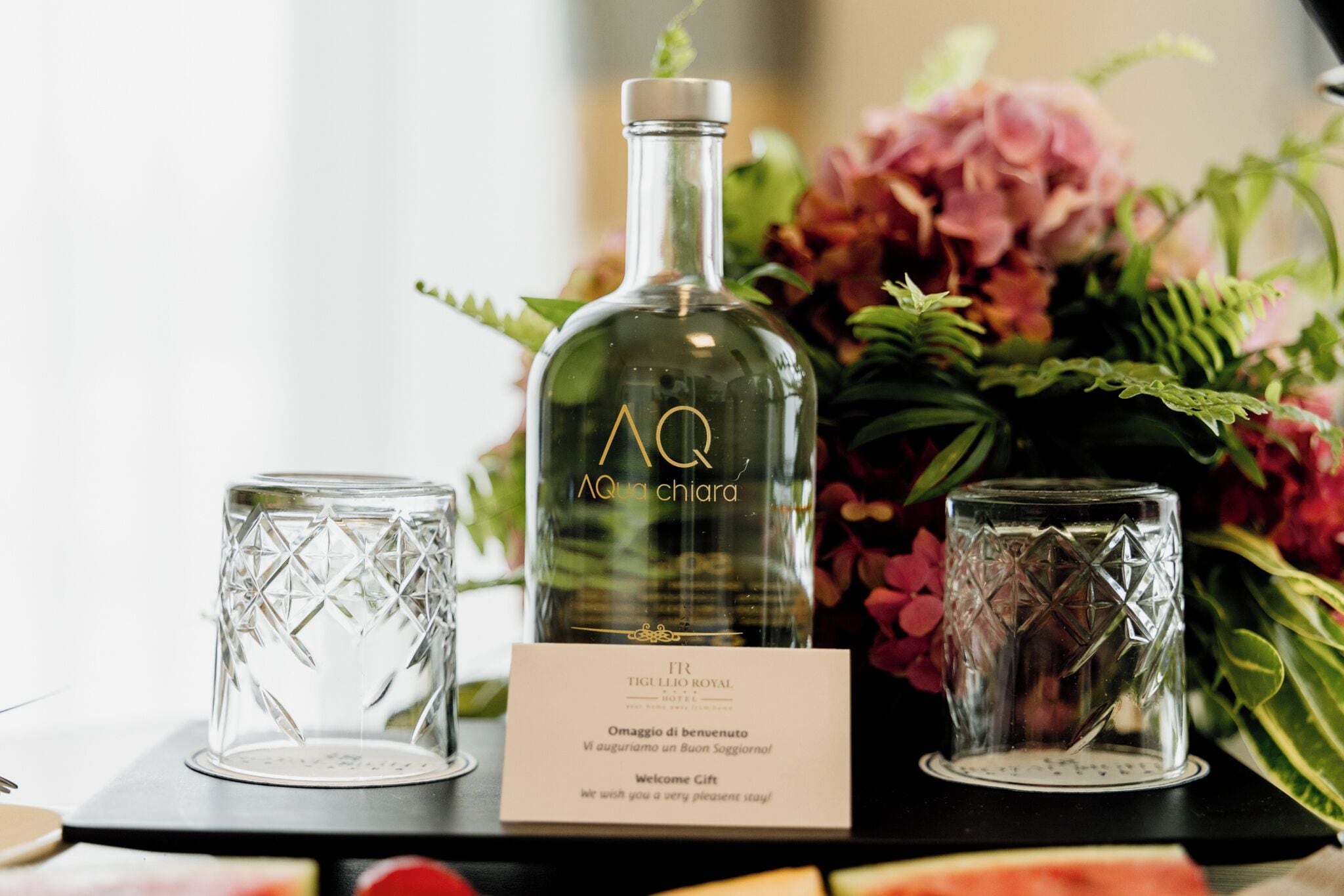 Eine Flasche AQ Aquaria, ein Glas mit geometrischem Muster und ein Glas mit Sternenmuster, alles auf einer schwarzen Oberfläche mit einem floralen Arrangement im Hintergrund.