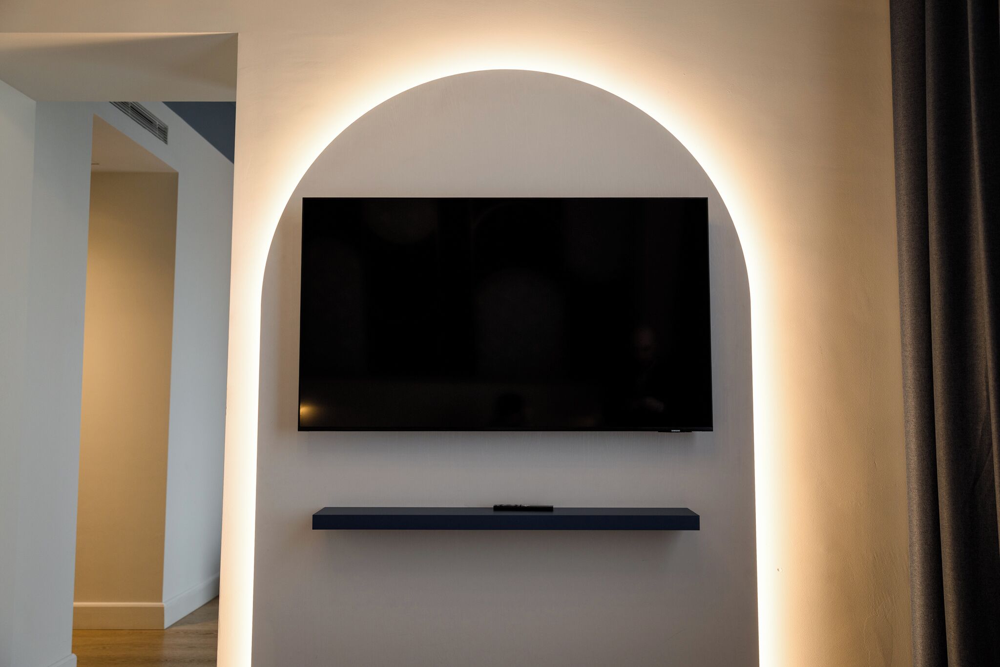 Un intérieur moderne avec un téléviseur à écran plat fixé sur un mur blanc, une étagère sombre en dessous et une porte donnant sur une autre pièce.