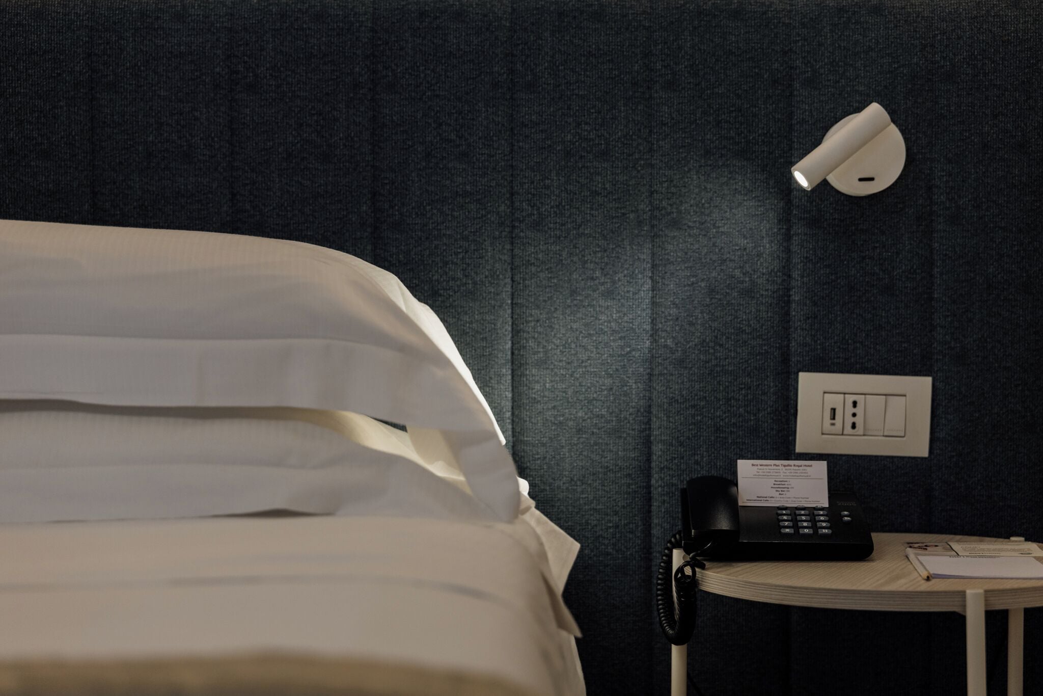 A hotel room with a bed, a bedside telephone, a lamp, and a small table with a book and papers.