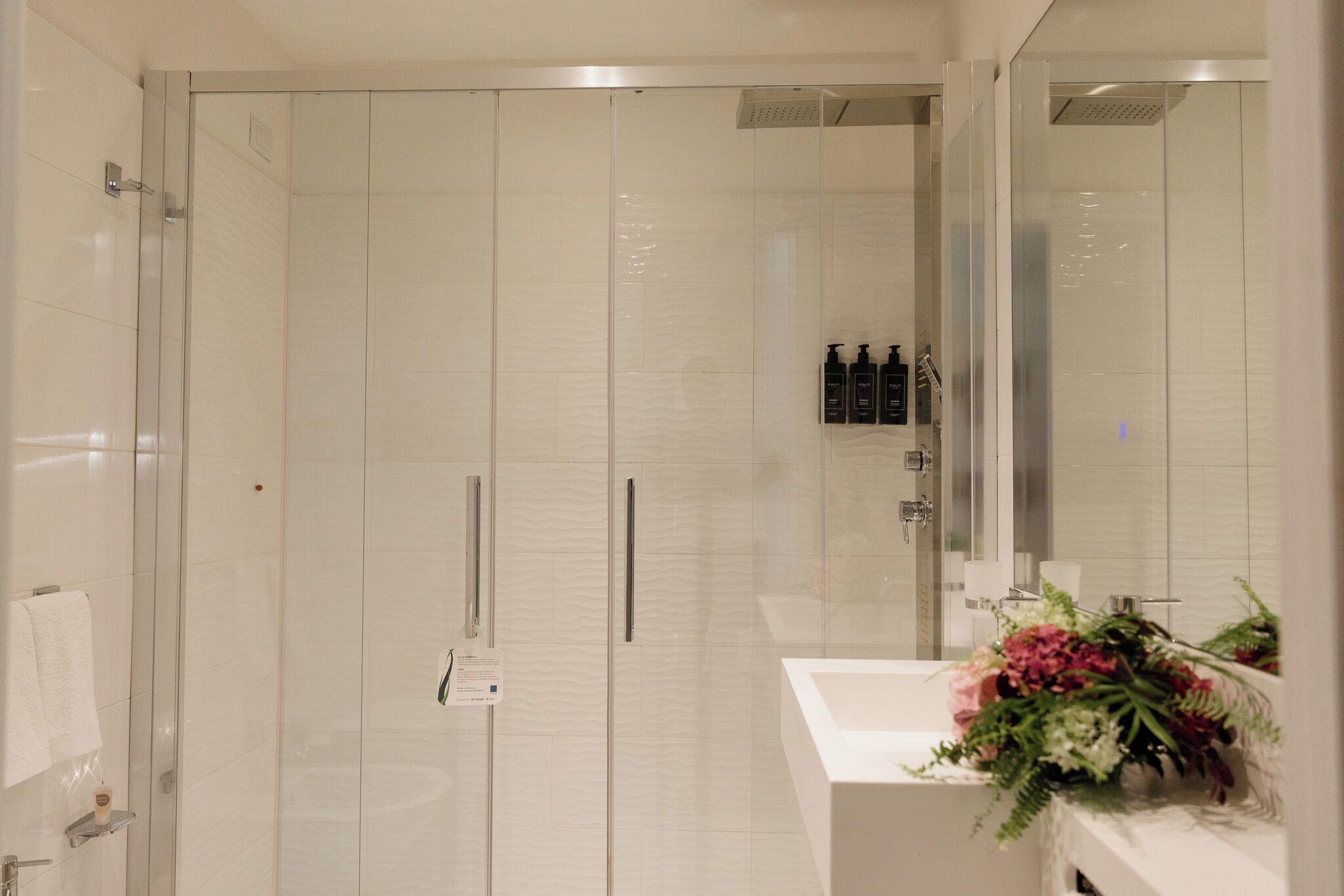 A modern bathroom with a glass shower enclosure, a white sink, and a mirror with a floral arrangement.