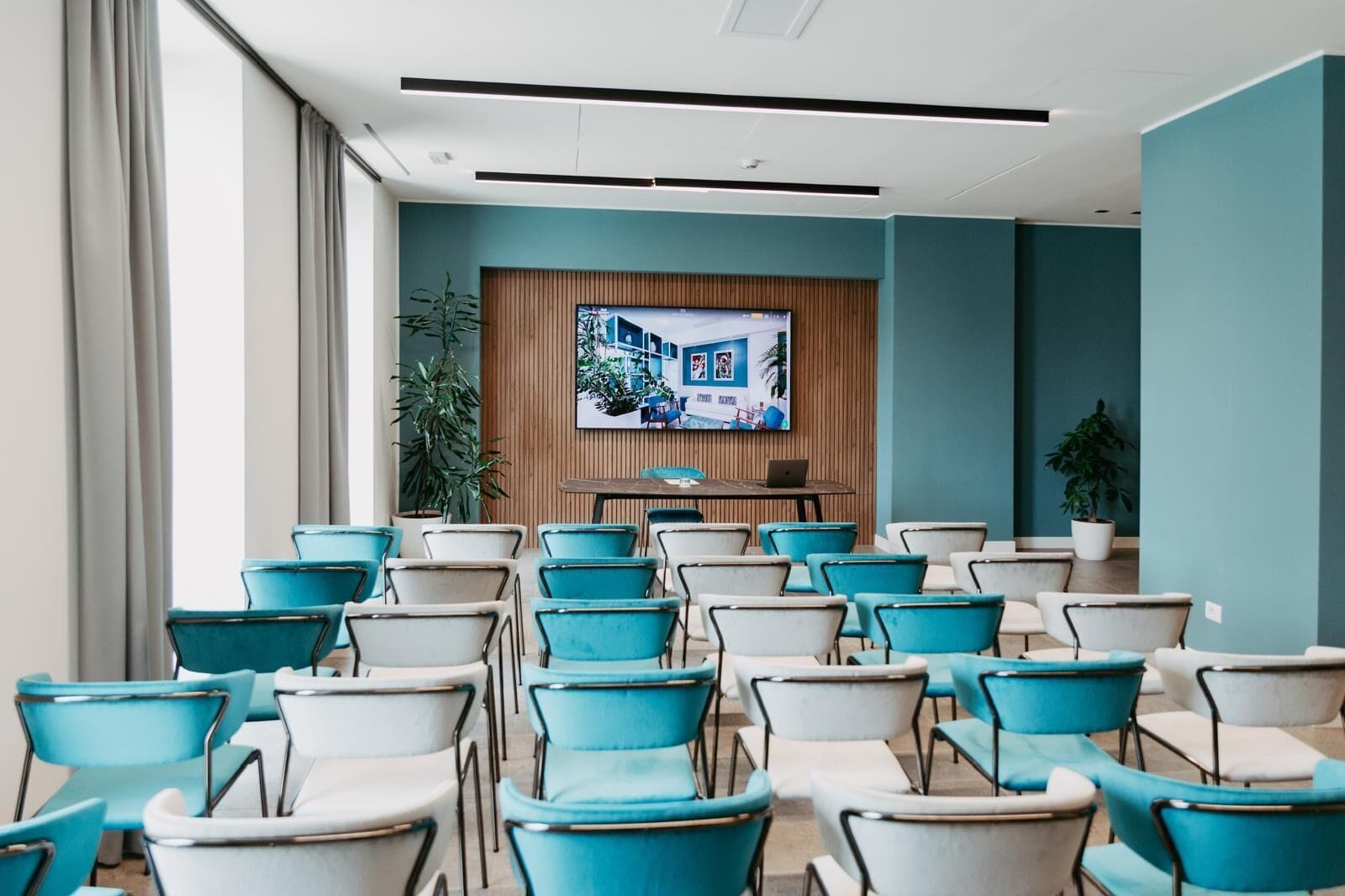 A conference room with rows of teal and white chairs, a wooden panel wall with a mounted screen, and potted plants.
