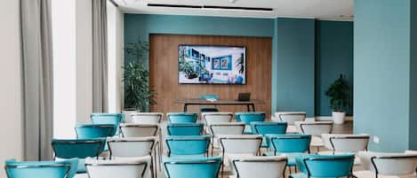 A conference room with rows of teal and white chairs, a wooden panel wall with a mounted screen, and potted plants.