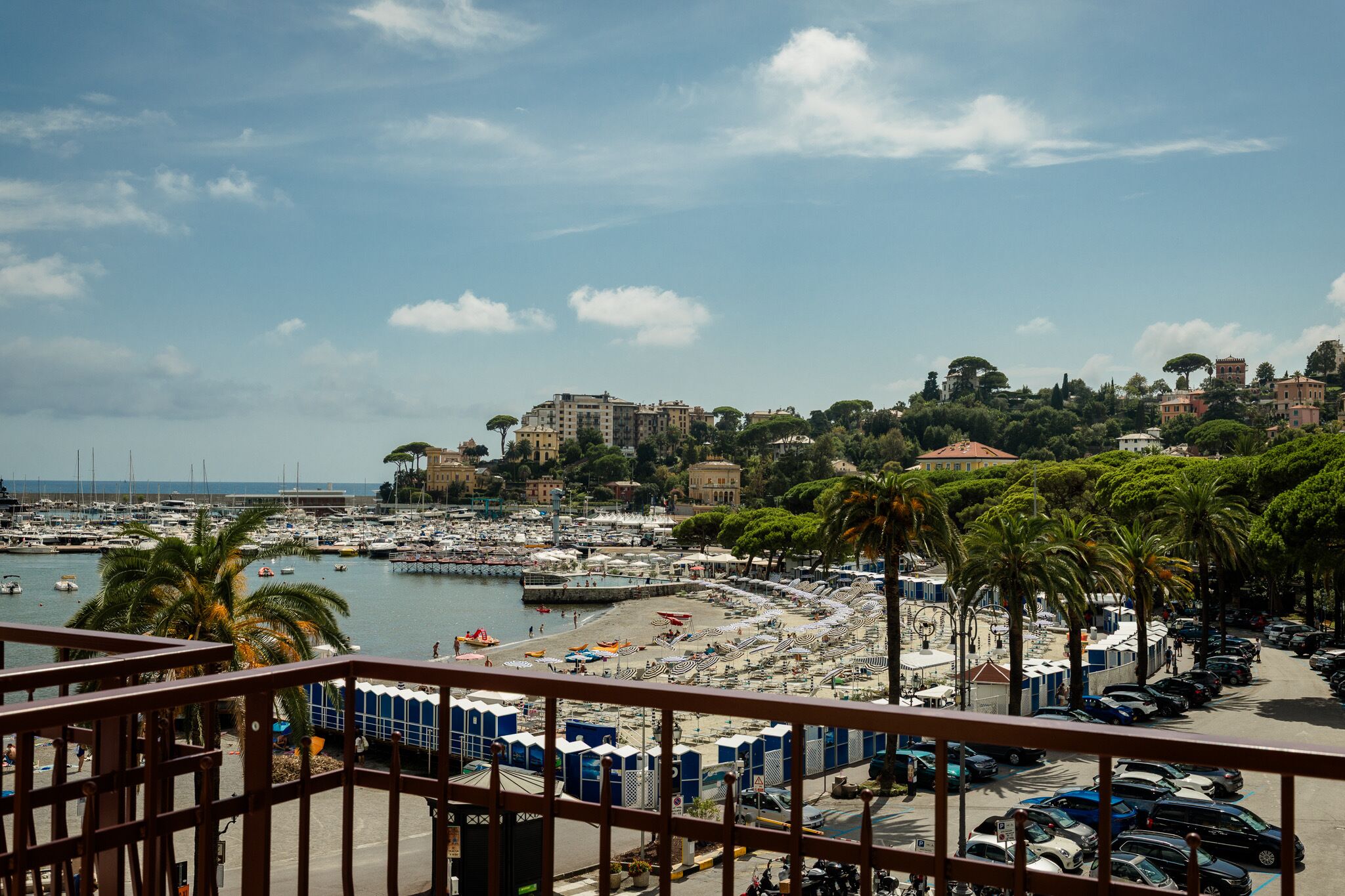 A coastal marina with boats, a beach with umbrellas, and buildings in the background.