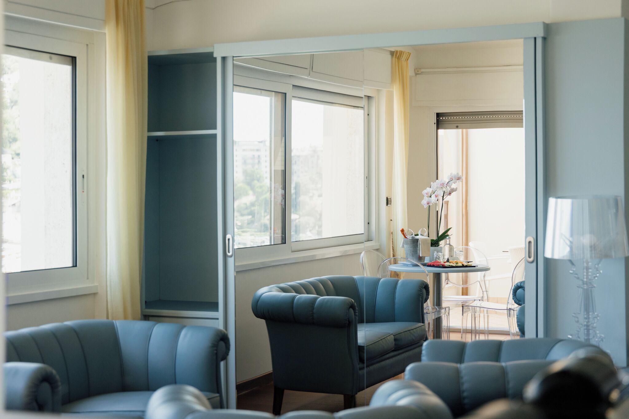 A room with a large window, a seating area with blue chairs, a table with a vase of flowers, and a mirror.