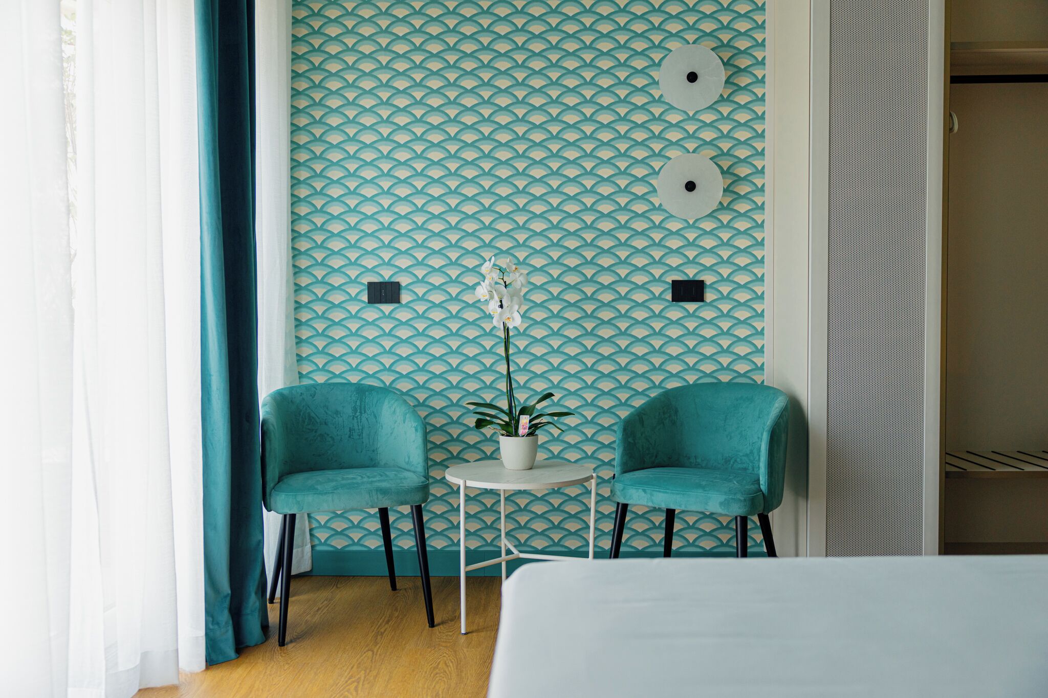 A room with teal patterned wallpaper, two teal chairs, a small round table with a potted plant, and a bed in the foreground.