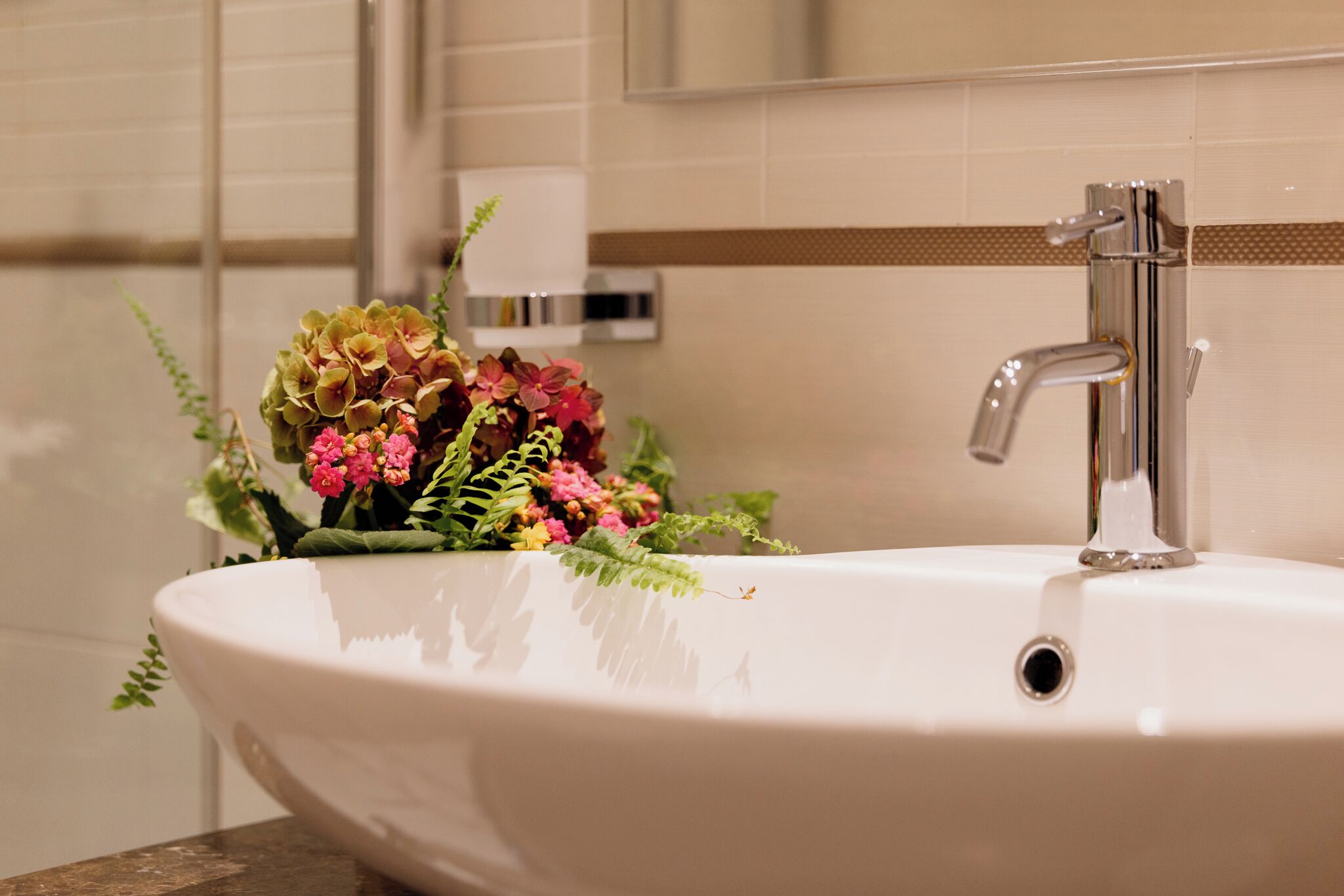 A bathroom sink with a modern faucet, a flower arrangement, and a towel dispenser in the background.