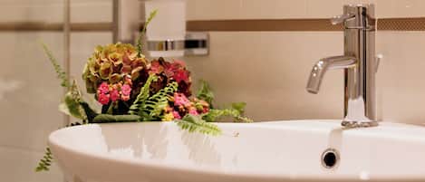 A bathroom sink with a modern faucet, a flower arrangement, and a towel dispenser in the background.
