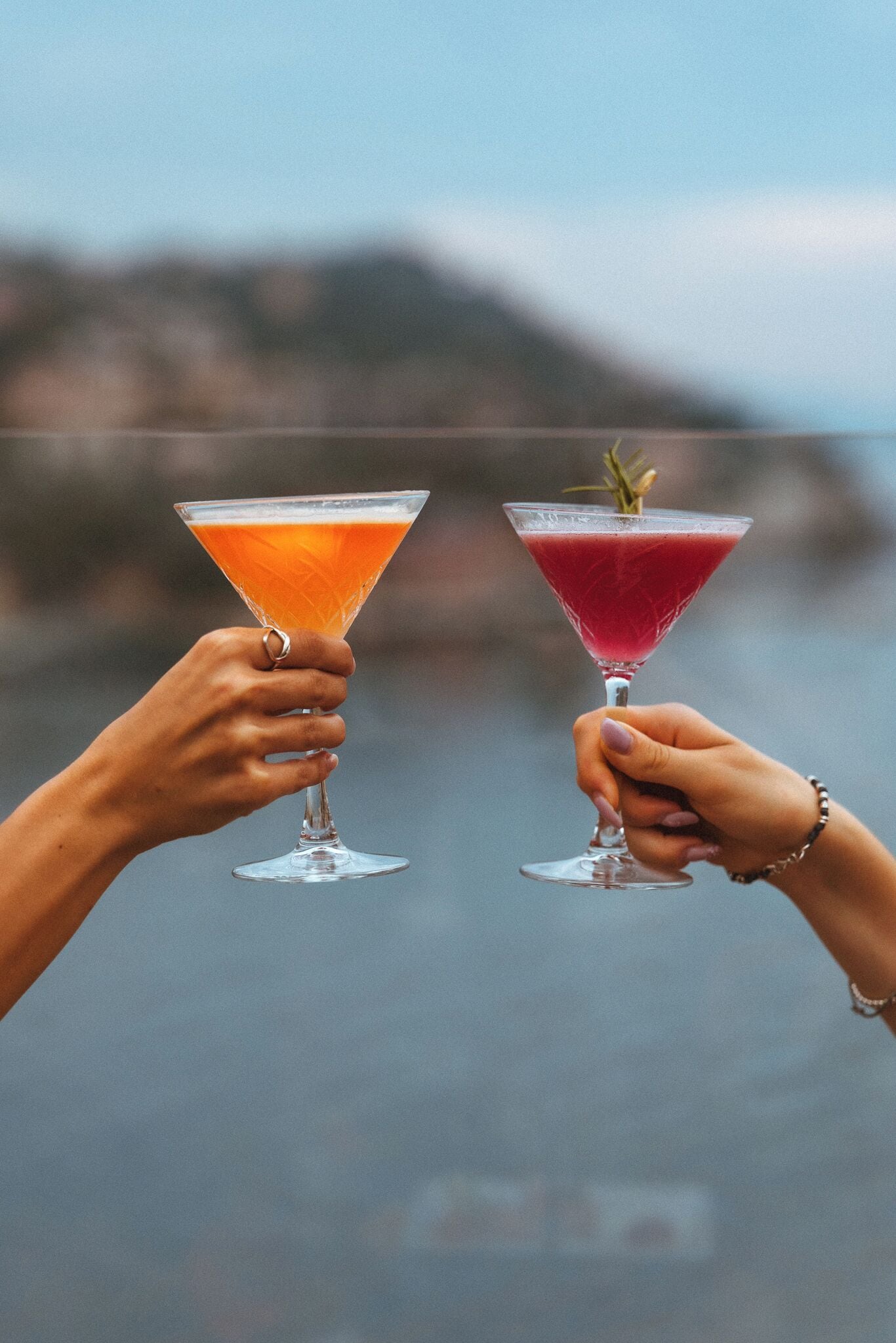 Two hands holding martini glasses with cocktails, one orange and one red, against a blurred background.