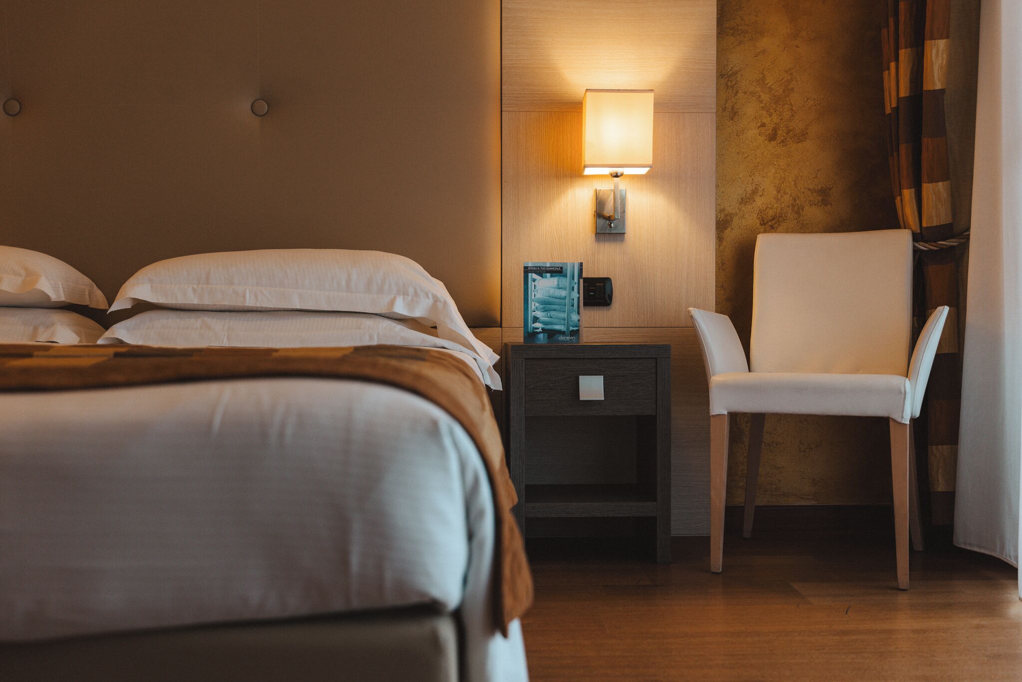A hotel room with a bed, a chair, a nightstand with a lamp, and a wall-mounted control panel.